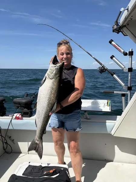 An angler on a charter boat holds up a large salmon, with rods set behind her and open water all around