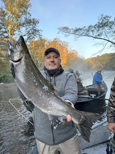 An angler in a drift boat holds up a large salmon on a misty river, with fall trees and other boats behind